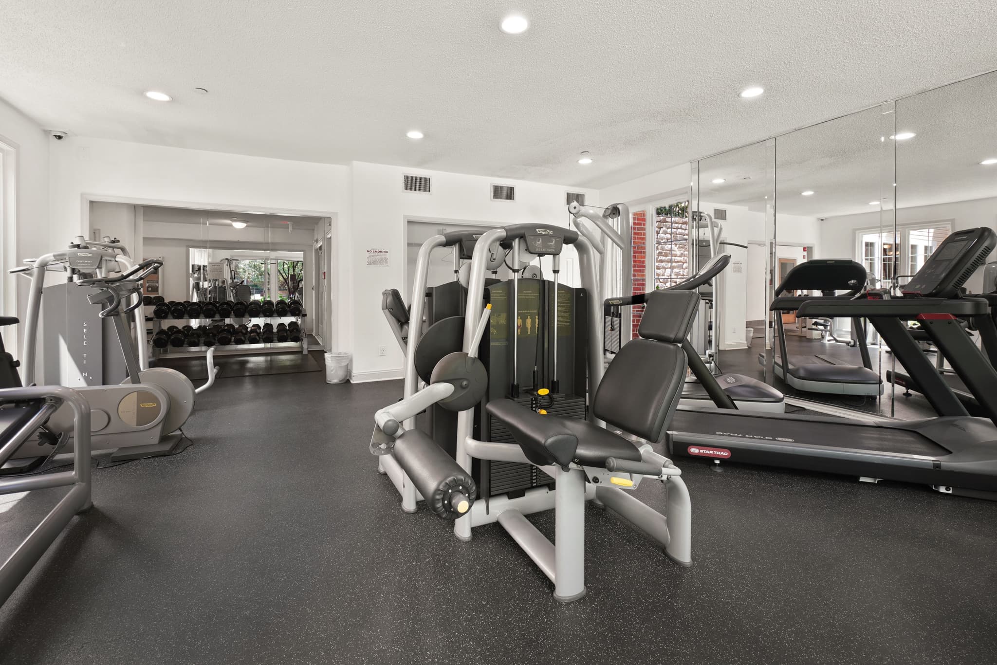 Helle, moderne Trainingsfläche eines Fitnessstudios mit Laufbändern, Kraftgeräten und Hantelständer im menschenleeren Zustand.