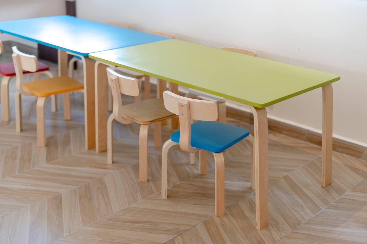 Heller Gruppenraum in einer Kindertagesstätte mit bunten Kindertischen und Holzstühlen auf glänzendem Parkettboden.