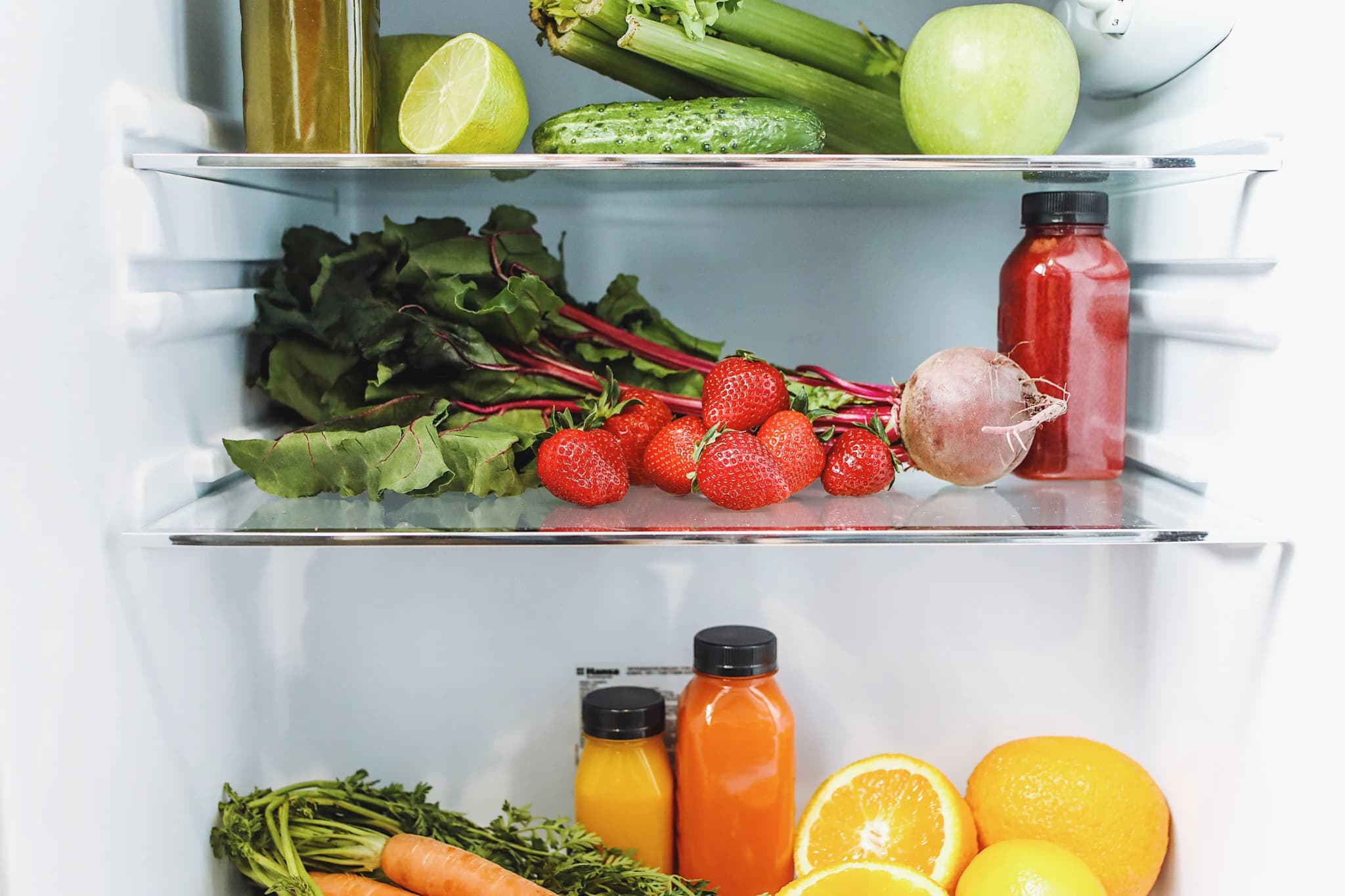 Geöffneter Kühlschrank mit Glasböden voller Erdbeeren, Mangold, Gurken, Orangen und Säften