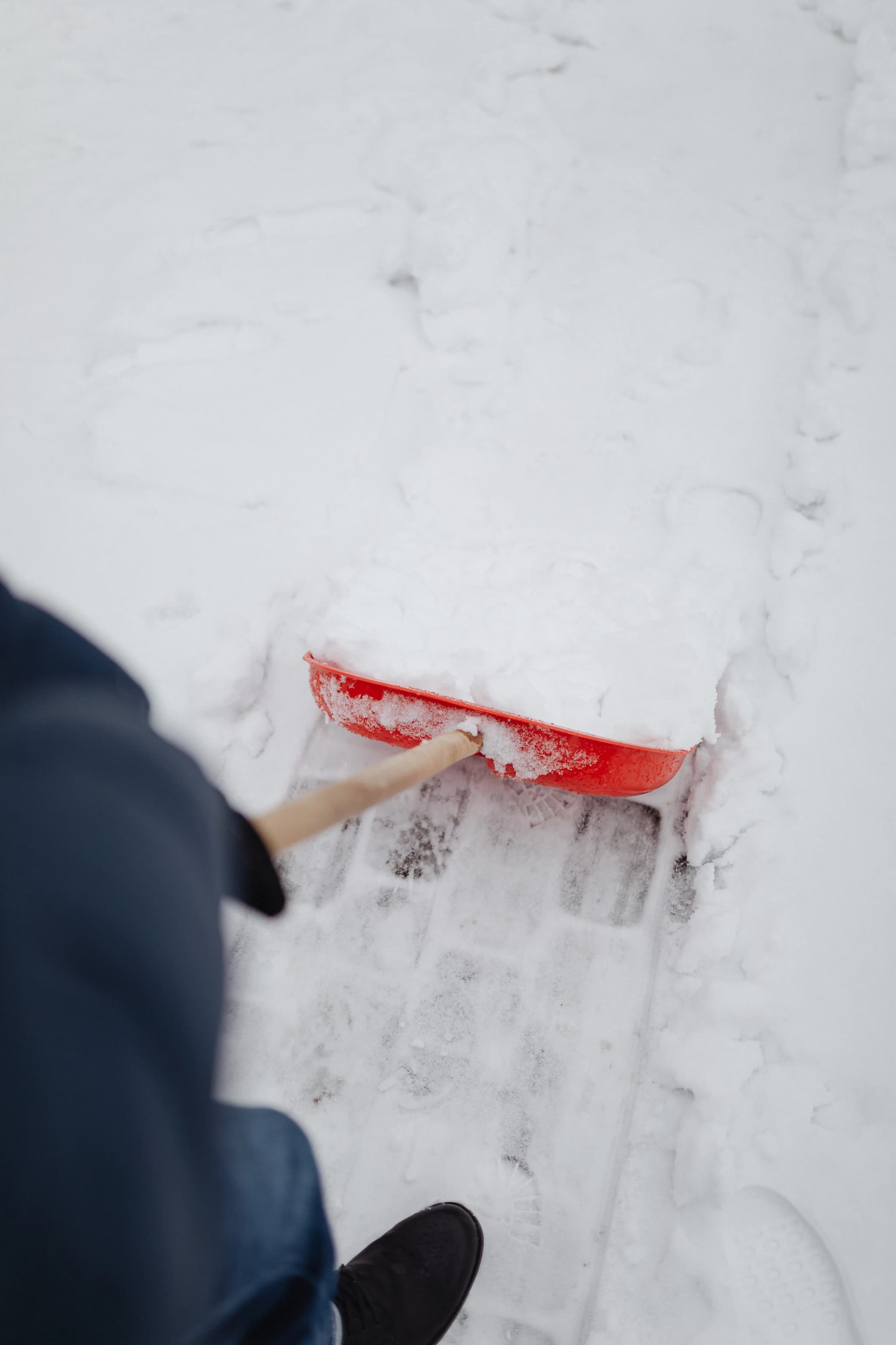 Person räumt mit einer roten Schneeschaufel Schnee von einem gepflasterten Gehweg